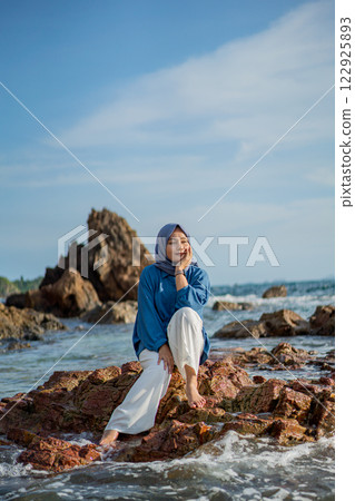Pensive Hijabi Woman Relaxing on Rocky Beach Enjoying Ocean Views and Coastal Scenery 122925893