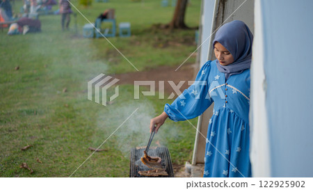 Young Muslim Woman Grilling Meat Outdoors at Picnic in a Beautiful Dress and Hijab Young Muslim Woman Grilling Meat Outdoors at Picnic in a Beautiful Dress and Hijab 122925902