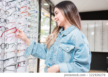 Young female buyer choosing glasses in an optical store. Customer choosing glasses in an eyewear store, Beautiful girl choosing glasses in an optical store 122926278