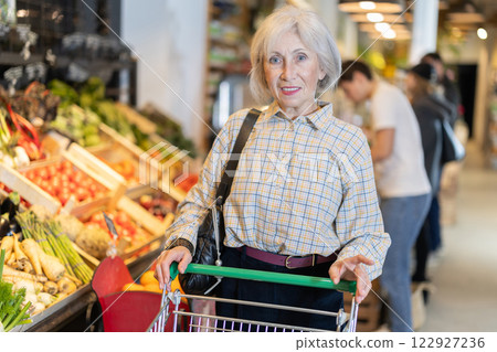 Elderly woman with shopping cart chooses groceries Elderly woman with shopping cart chooses groceries 122927236