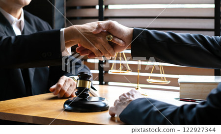 Close-up of two business professionals shaking hands over wooden desk with gavel, scales of justice, and legal books, agreement, legal consultation, or business negotiation Close-up of two business professionals shaking hands over wooden desk with gavel, scales of justice, and legal books, agreement, legal consultation, or business negotiation 122927344