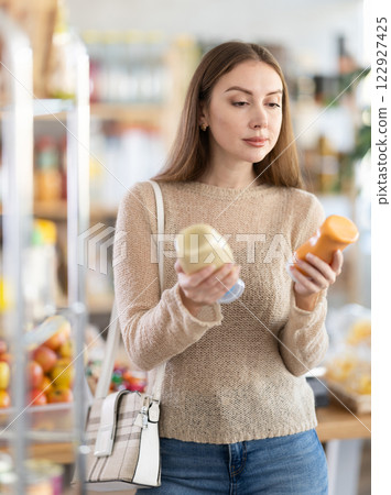 Smiling young female shopper carefully examining mayonnaise on shelves of food store, choosing flavorful addition to dinner 122927425