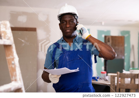 Foreman in blue overalls and protective helmet is talking on mobile phone in renovated room Foreman in blue overalls and protective helmet is talking on mobile phone in renovated room 122927473