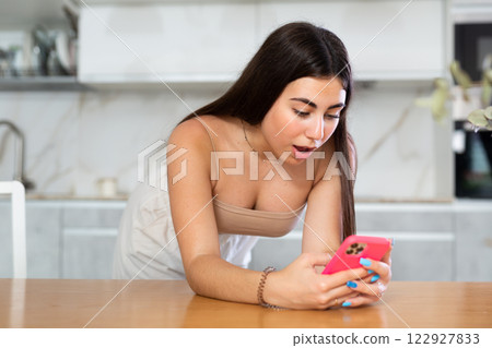 Young woman leaning on kitchen table and looking at her phone Young woman leaning on kitchen table and looking at her phone 122927833