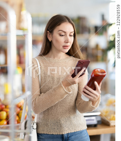 Interested young girl pondering choice of healthy and tasty ketchup, scanning barcode with smartphone to get information about product in grocery store Interested young girl pondering choice of healthy and tasty ketchup, scanning barcode with smartphone to get information about product in grocery store 122928559