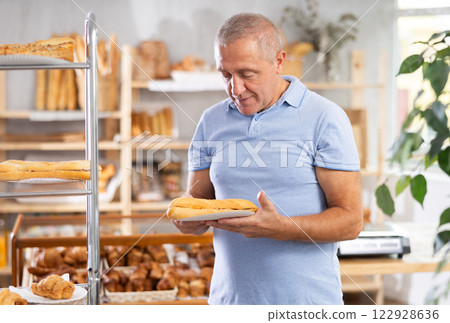 Senior male customer stands with croissant in hands near window of bakery. 122928636