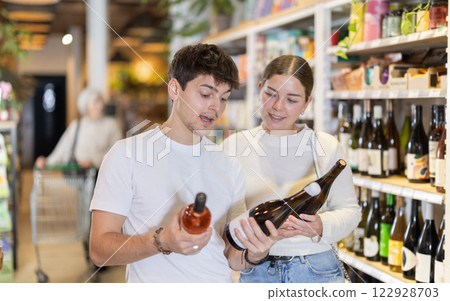 Guy and girl spouses customers in supermarket examines bottles of wine, chooses alcoholic drink. 122928703