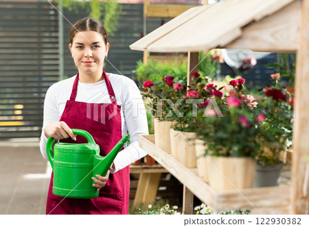 employee of flower greenhouse stands with watering can near shelf with plants 122930382