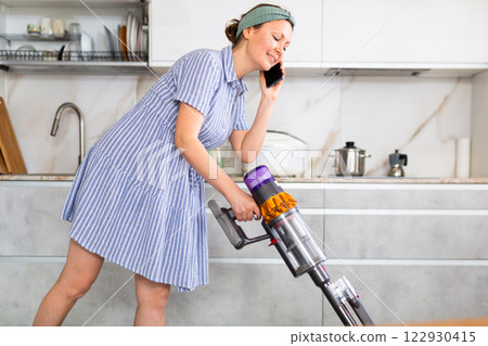 Woman in dress and headband is vacuuming floor and talking on mobile phone, ordering food delivery Woman in dress and headband is vacuuming floor and talking on mobile phone, ordering food delivery 122930415