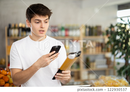 In shop young man scans QR code and take reading on spaghetti package using phone camera In shop young man scans QR code and take reading on spaghetti package using phone camera 122930537