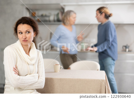 Frustrated woman sitting in kitchen during quarrel between her husband and elderly mother. 122930640