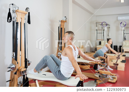 Young man stretching his arms using pilates reformer while sitting backwards 122930973