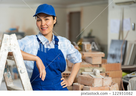 Portrait of positive builder woman in blue overalls next to stepladder Portrait of positive builder woman in blue overalls next to stepladder 122931082
