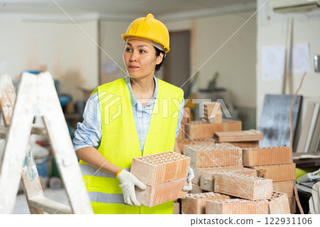 Female builder in a yellow vest and hardhat carrying hollow clay bricks and putting them on brick stack Female builder in a yellow vest and hardhat carrying hollow clay bricks and putting them on brick stack 122931106