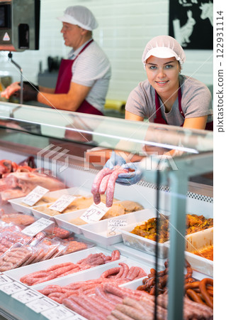 Young saleswoman arranging uncooked butifarra sausages in glass showcase 122931114