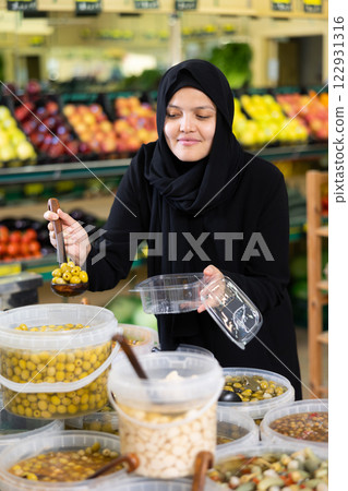 Asian woman in dark blue hijab puts handful of olives in disposable container with ladle Asian woman in dark blue hijab puts handful of olives in disposable container with ladle 122931316