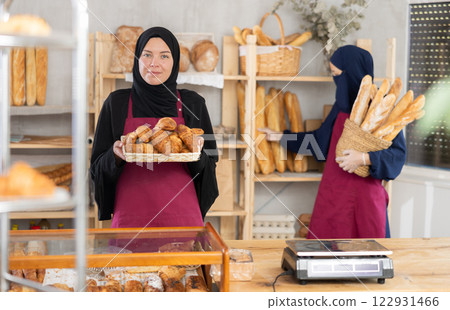 Young Muslim saleswoman demonstrates croissants while another woman stocks baguettes Young Muslim saleswoman demonstrates croissants while another woman stocks baguettes 122931466