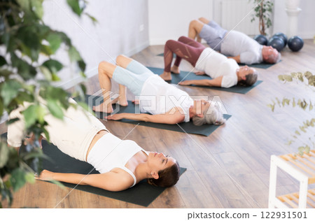 Class of mature women stretching gluteal muscle while lying on back during yoga Pilates workout on mats in gym studio Class of mature women stretching gluteal muscle while lying on back during yoga Pilates workout on mats in gym studio 122931501