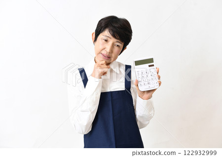 A middle-aged woman wearing an apron with a troubled look on her face and holding a calculator A middle-aged woman wearing an apron with a troubled look on her face and holding a calculator 122932999