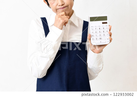 一名中年婦女身穿圍裙，帶著憂慮，手裡拿著計算器 122933002