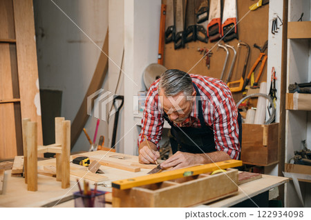 A carpenter in a plaid shirt carefully measures wooden pieces on workbench using a framing square, ensuring precision in each cut. The workshop is filled with tools and materials for furniture making 122934098