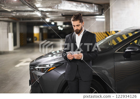 Young Caucasian male businessman counts cash in underground parking lot next to car, conveying secrecy and business. 122934126