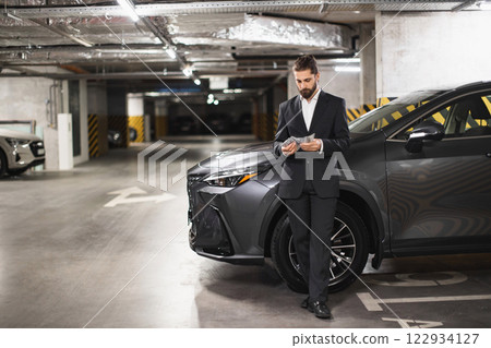 Caucasian male businessman in suit counts cash by car in underground parking lot. Scene conveys secrecy, finance, and wealth in urban setting. 122934127