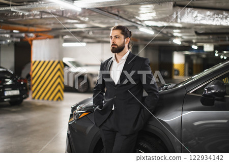 Young Caucasian man in formal suit leaning against car in underground parking lot. Business professional exuding confidence, poised in urban setting. Modern lifestyle and transportation theme. Young Caucasian man in formal suit leaning against car in underground parking lot. Business professional exuding confidence, poised in urban setting. Modern lifestyle and transportation theme. 122934142