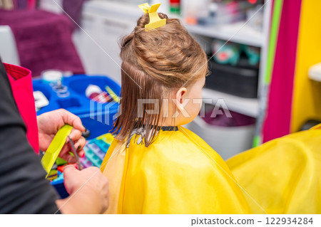 Back view of hairdresser making a hair style to cute little girl in real child salon with colorful interior and toys 122934284
