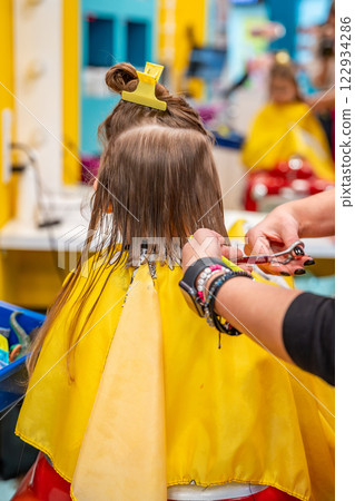 Back view of hairdresser making a hair style to cute little girl in real child salon with colorful interior and toys 122934286