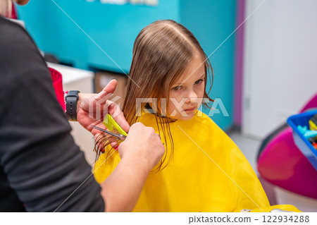 Hairdresser making a hair style to cute little girl in real child salon with colorful interior and toys 122934288