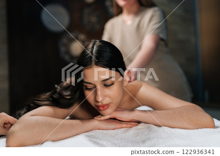 Closeup portrait of tranquil young woman lying on massage table with closed eyes during professional relaxing massage at spa salon. Professional massage therapist performing back treatment. Closeup portrait of tranquil young woman lying on massage table with closed eyes during professional relaxing massage at spa salon. Professional massage therapist performing back treatment. 122936341