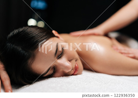 Close-up face of tranquil young woman client relaxing during back massage in spa salon with dark interior. Professional massage therapist providing therapeutic back massage to relaxed female. Close-up face of tranquil young woman client relaxing during back massage in spa salon with dark interior. Professional massage therapist providing therapeutic back massage to relaxed female. 122936350