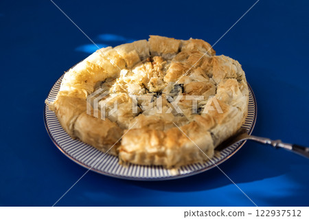 A close-up of a plate with a delicious Greek pie. Tasty spanakopita with spinach and cheese on a blue table. A close-up of a plate with a delicious Greek pie. Tasty spanakopita with spinach and cheese on a blue table. 122937512