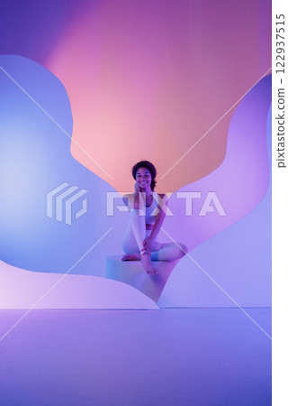 Young African is sitting cross-legged on round platform. Black athletic girl in white fitness suit smiles 122937515