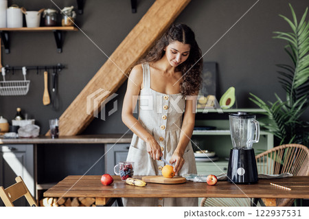 Charming curly-haired brunette girl in linen sundress prepares fruit smoothie in blender in kitchen. Charming curly-haired brunette girl in linen sundress prepares fruit smoothie in blender in kitchen. 122937531