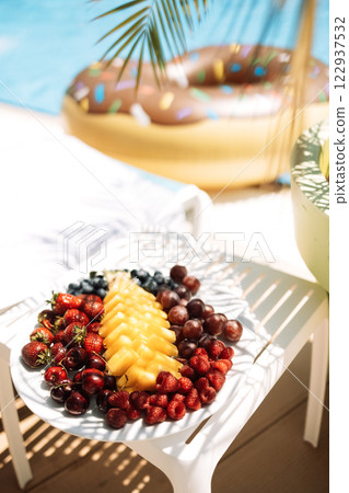 Delicious juicy fruit and berry set on the table by the pool. Pineapple, cherries, strawberries, raspberries and grapes on a plate next to the chaise longue. Tasty snacks for lunch. Delicious juicy fruit and berry set on the table by the pool. Pineapple, cherries, strawberries, raspberries and grapes on a plate next to the chaise longue. Tasty snacks for lunch. 122937532