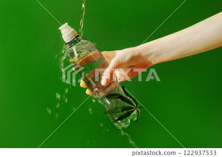 A close-up of a female hand with a neat manicure holding a mock-up plastic bottle with a cannabis leaf. Fresh mineral water with splashes and hemp. 122937533