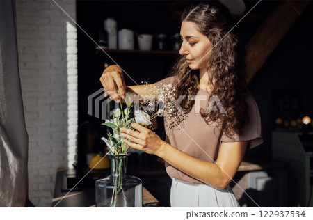 Charming curly-haired brunette in casual clothes collects bouquet in dining room. Young attractive woman puts flowers in glass vase. Cute girl decorates her house with flowers. Charming curly-haired brunette in casual clothes collects bouquet in dining room. Young attractive woman puts flowers in glass vase. Cute girl decorates her house with flowers. 122937534