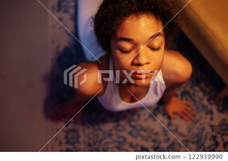 An attractive African practices evening yoga. A charming curly black girl in a fitness suit stretches her back on the floor in the living room. Top view. An attractive African practices evening yoga. A charming curly black girl in a fitness suit stretches her back on the floor in the living room. Top view. 122939990