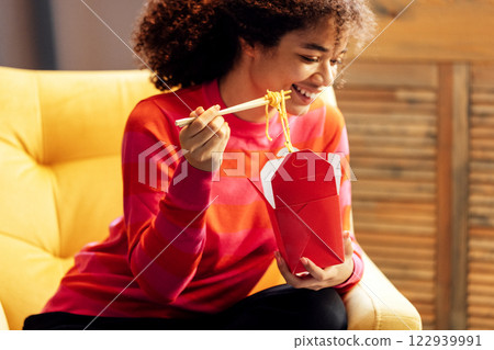 Cute African woman in sweater is sitting in yellow chair and eating noodles with chopsticks Cute African woman in sweater is sitting in yellow chair and eating noodles with chopsticks 122939991