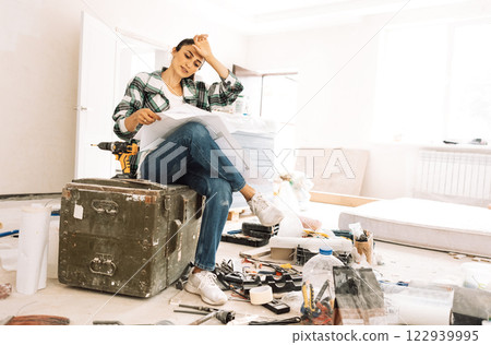 Young tired woman with house plan is sitting on box among building materials and inventory. Girl in casual clothes is thinking about home renovation. Young tired woman with house plan is sitting on box among building materials and inventory. Girl in casual clothes is thinking about home renovation. 122939995