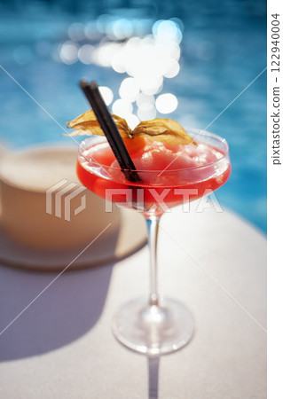 A red drink in an elegant glass with a long stem on the side of the pool. An alcoholic ice drink decorated with a flower on a background of turquoise water. A red drink in an elegant glass with a long stem on the side of the pool. An alcoholic ice drink decorated with a flower on a background of turquoise water. 122940004