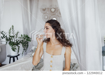A charming curly-haired brunette in a linen sundress holds a transparent cup and drinks water. 122940012