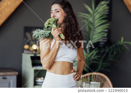 An attractive smiling vegan girl in a white top bites fresh broccoli. An attractive smiling vegan girl in a white top bites fresh broccoli. 122940015