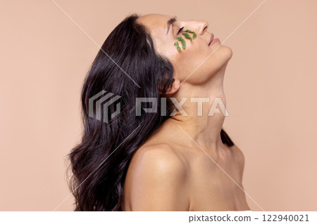 Studio portrait of a young woman with leaves under her eye on a beige Studio portrait of a young woman with leaves under her eye on a beige 122940021