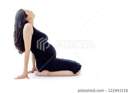 A young attractive pregnant woman in a black dress in profile. The charming mother-to-be is sitting on the floor. White isolated background. A lady expecting a baby. A young attractive pregnant woman in a black dress in profile. The charming mother-to-be is sitting on the floor. White isolated background. A lady expecting a baby. 122941516