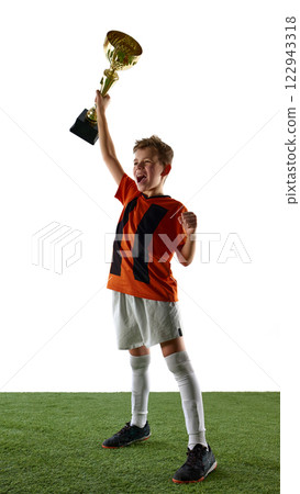 Full-length image of boy, soccer player in orange jersey and white shorts, standing on grassy surface, lifting trophy. Victory Full-length image of boy, soccer player in orange jersey and white shorts, standing on grassy surface, lifting trophy. Victory 122943318
