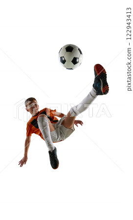 Dynamic image of boy, soccer player in orange jersey and white shorts kicking ball with extended leg, showing skill against white studio background. 122943413