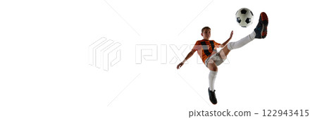 Boy in orange jersey, white shorts, white socks, airborne, kicking soccer ball with extended leg, showing skill against white studio background. 122943415